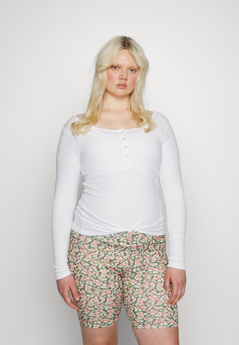 Blusa branca de mangas compridas em ribana com um decote abotoado, combinada com calções verdes estampados com flores em tons de rosa e branco.