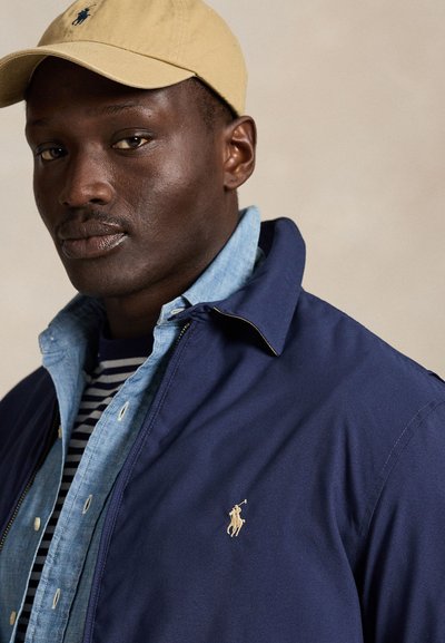 Mann mit beiger Kappe, dunkelblauer Jacke mit Polo-Logo, hellblauem Hemd und dunkelblau-weiß gestreiftem Hemd, der leicht seitlich vor einem einfarbigen Hintergrund schaut.