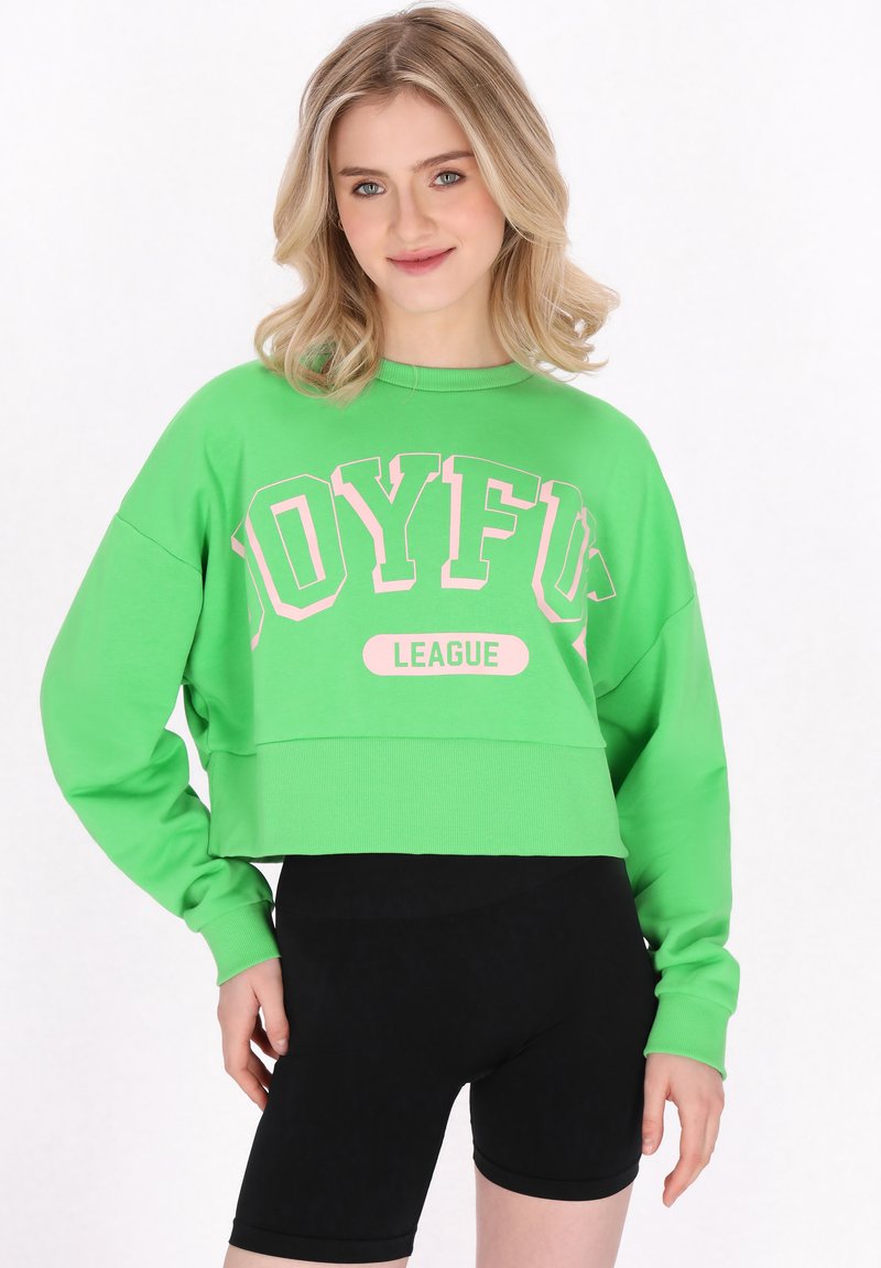 Zöld, rövid cropped pulóver, pink "JOYFUL LEAGUE" felirattal. Hosszú ujja van és kerek nyakú. Fekete kerékpáros sortokkal kombinálva.