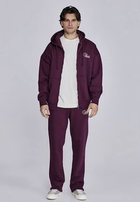 Vinröd sweatshirt med huva och fickor, kombinerad med matchande sweatpants. Har vita logotypdetaljer och är tillverkad av mjukt tyg.