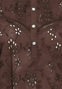 Top in tessuto marrone con ricamo floreale intricato in marrone scuro e dettagli ritagliati. Chiusura frontale con bottoni bianchi.