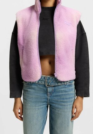 Person trägt eine hellviolette Fleeceweste über einem schwarzen, kurz geschnittenen Langarmshirt, kombiniert mit blauen Jeans und einem Gürtel.