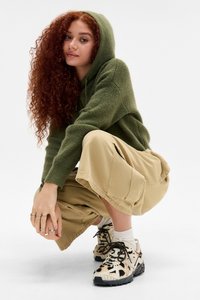 Sudadera verde de punto, pantalones cortos de cargo beige, zapatos estampados con detalles negros. Pose en cuclillas, fondo claro.
