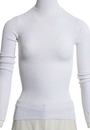 Pull col roulé blanc à manches longues avec motifs géométriques perforés, porté sur un mannequin avec un pantalon clair.