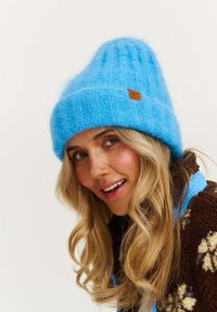 Svetlomodrá pletená čiapka s textúrovaným vzorom, ktorá je ozdobená obdĺžnikovou hnedou koženou štítkom, nosená s hnedou flísovou bundou.