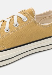 Lærredssneakers i sennepsgul med hvite snører, metalløyler og en kontrasterende svart stripe på sålen. Strukturerte tekstiloverflater.