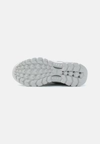 Semelle de chaussure de sport blanche avec des rainures profondes et angulaires pour une meilleure adhérence. Surface texturée avec des motifs prononcés et une marque intégrée dans le caoutchouc.