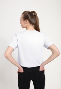 Žena s hnědými vlasy v culíku, oblečená v bílé tričko a černých džínách, s rukama v zadních kapsách, stojící bokem proti jednoduchému pozadí.