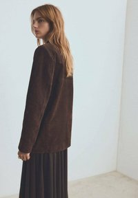 Giacca in suede marrone caratterizzata da un design rilassato, abbinata a una gonna a pieghe scura e fluttuante. Texture liscia, con dettagli hardware minimi.