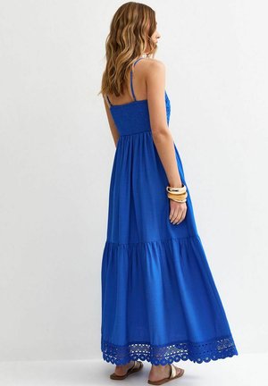 Mujer con cabello ondulado, vestida con un vestido maxi azul sin mangas con borde de encaje, brazaletes dorados y sandalias planas, de pie contra un fondo claro y liso.