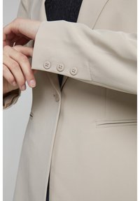 Blazer beige con un design sartorial, texture liscia e quattro bottoni coordinati sui polsini. Include un sottile dettaglio della tasca.