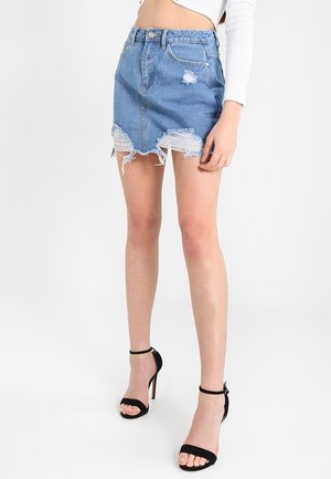 Femme portant une mini-jupe en jean bleu délavé, un top blanc court à manches longues et des sandales noires à talons hauts avec bride à la cheville.