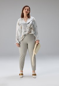 Wit en beige blouse met marmerpatroon en een keyhole-halslijn, gecombineerd met lichtgrijze strakke broek en beige handtas. Bruine en witte platte schoenen.
