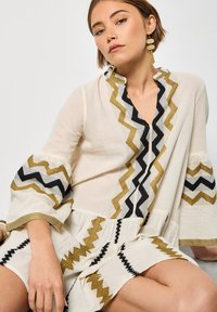 Robe tunique blanche avec broderies en zigzag dorées, noires et blanches. Comprend des manches évasées et un décolleté en V. Tissu léger avec une coupe décontractée.