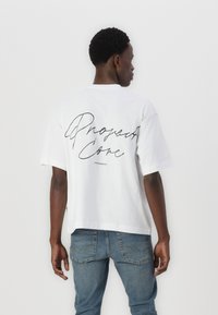 White cotton T-shirt with a loose fit, featuring a black cursive "Project Core" print on the back and short sleeves.