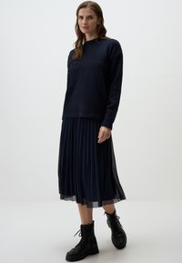 Pull en maille marine avec un design brodé, associé à une jupe midi plissée marine et des bottes de combat noires à lacets. Texture lisse et look superposé.