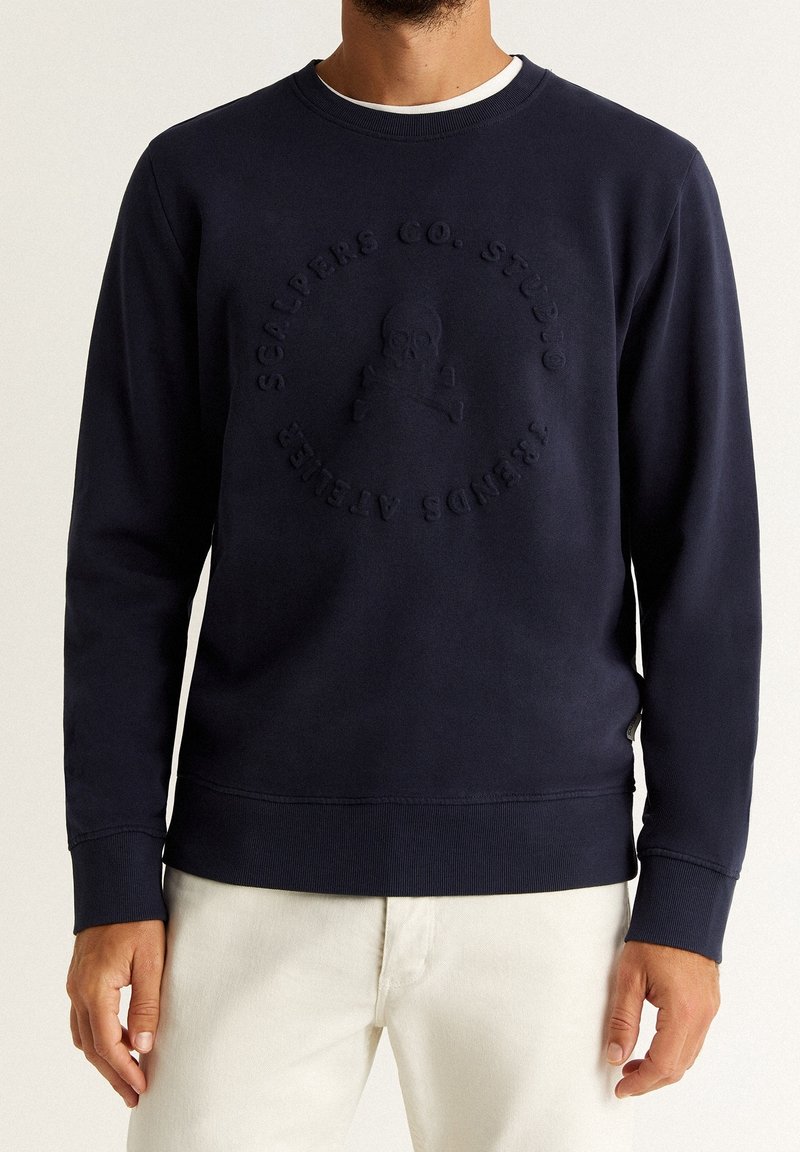 Homme portant un sweat-shirt à col rond bleu marine avec un logo en relief de crâne et os croisés ainsi qu'un texte circulaire à l'avant, assorti d'un pantalon beige clair.
