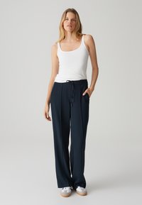 Pantaloni a gamba larga blu navy con vita elastica, tasche laterali e una consistenza liscia, abbinati a una canotta bianca e sneakers.