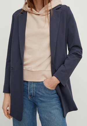 Femme portant un blazer bleu marine par-dessus un sweat à capuche beige, une main dans la poche d’un jean bleu clair, sur un fond uni.