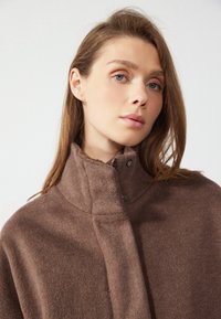 Veste en polaire marron avec un col haut, dotée d'une fermeture à pression et d'une texture douce, présentée sur un fond neutre.