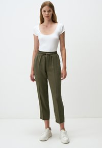 Haut blanc à manches courtes côtelé associé à un pantalon tapered olive vert, à taille élastique. Le modèle porte des baskets blanches. Fond uni.