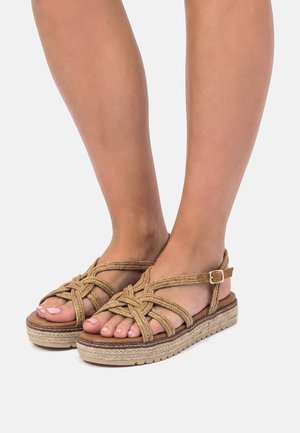 Füße, die sandfarbene Plateausandalen mit geflochtenen Riemen und Schnallenverschluss tragen, stehen vor einem schlichten hellen Hintergrund.