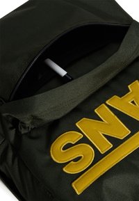 Zelený látkový batoh má velké žluté vyšívané logo "SNA", zapínání na zip a viditelný pero uvnitř.