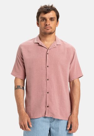 Camisa de manga corta en tela rosa claro con un acabado texturizado, con botones en la parte delantera y cuello, combinada con jeans de color azul claro.