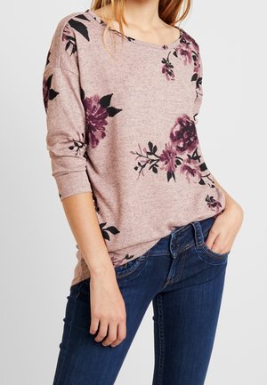 Woman wearing a long-sleeve pink sweater with purple and black floral patterns paired with dark blue jeans, hand in pocket.