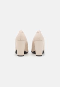 Chaussures à talons hauts beiges avec une texture lisse, un bout arrondi et un talon bloc. Le talon possède une base noire contrastante pour un détail supplémentaire.