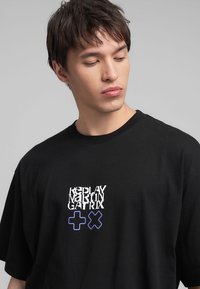 T-shirt nera in cotone con vestibilità oversize. Presenta una grafica stampata in bianco e blu sulla parte anteriore, inclusi simboli. Collo rotondo.
