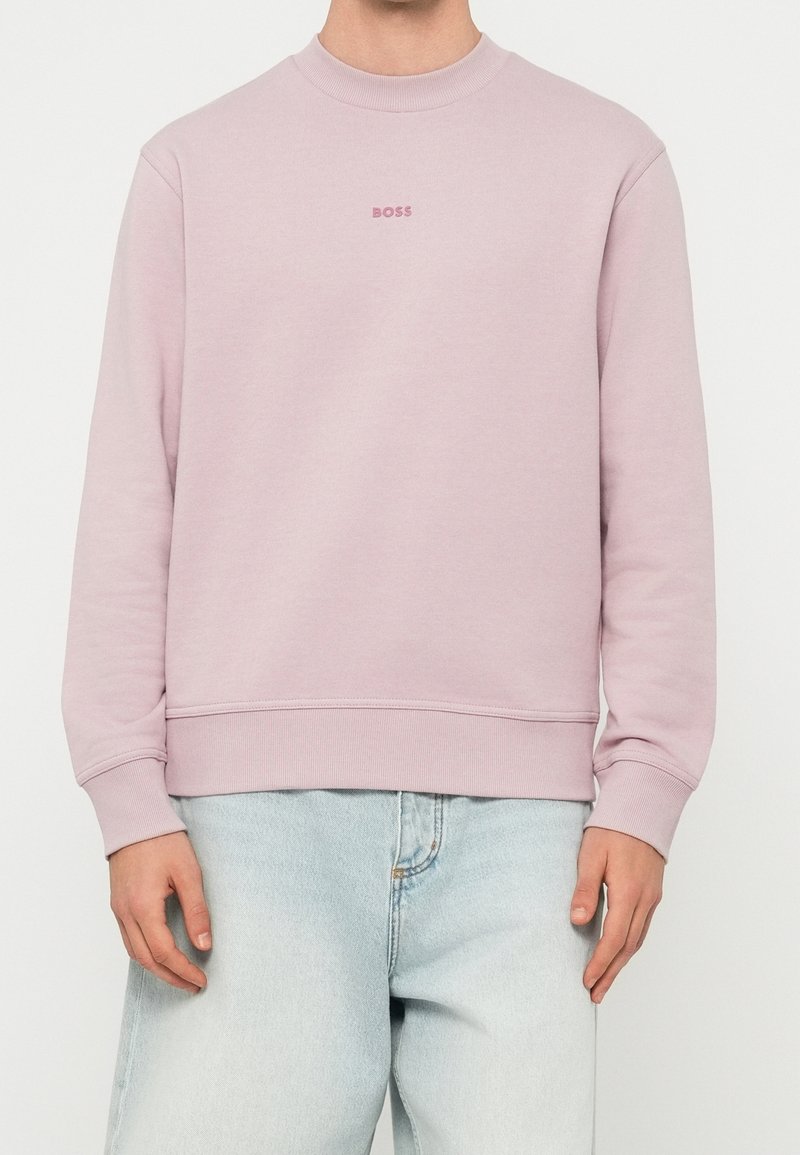 Personne portant un sweat-shirt rose clair à col rond avec le texte « BOSS » sur la poitrine et un jean bleu clair, debout, les mains détendues sur les côtés.