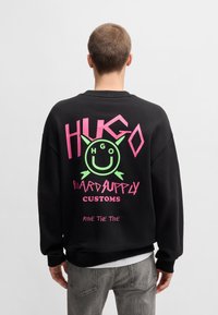 Felpa nera con grafica testuale vibrante in rosa e verde. Vestibilità ampia, scollatura tonda, con il design di 'HUGO' e una faccina sorridente sul retro.