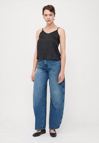 Schwarzes Satin-Cami mit dünnen Trägern, kombiniert mit blauen Jeans im weiten Bein. Frau trägt schwarze verziert Flache Schuhe und steht vor einem weißen Hintergrund.