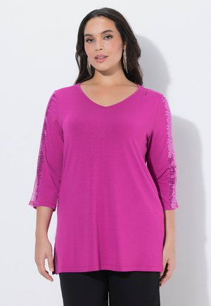 Kvinde iført en skrigende pink tunika med V-hals og 3/4-ærmer med lodrette pailletstriber, kombineret med sorte bukser og lange øreringe.