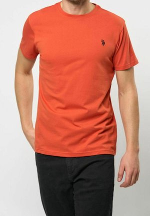 Mann trägt ein schlichtes orangefarbenes T-Shirt mit kurzen Ärmeln und einem kleinen schwarzen bestickten Logo auf der Brust, kombiniert mit schwarzen Hosen.