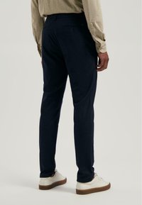 Man in een marineblauwe broek en witte sneakers, staande met zijn rug naar de camera tegen een effen lichte achtergrond.