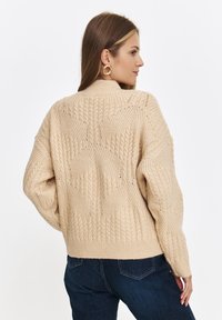 Beżowy sweter dziany z wzorem w warkocze, ściągacz na dole i wysoki kołnierz. Teksturowane wykończenie z siateczkowym detalem na plecach. Połączony z ciemnymi dżinsami.