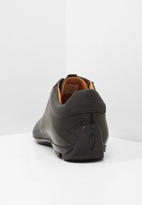 Chaussure en cuir noir avec design perforé, talon texturé et intérieur contrastant en cuir marron clair. Présente un détail de logo et un talon arrondi.
