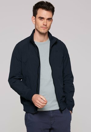 Navy lightweight jacket with a zip front, ribbed cuffs, and a streamlined design. Worn over a light gray shirt, features minimal branding.
