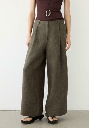 Kobieta nosząca szerokie oliwkowozielone spodnie, czarne sandały na obcasie, brązowy top bez rękawów oraz długi naszyjnik z owalnym wisiorkiem.