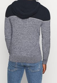 Mörkblå och grå stickad hoodie med texturerad kropp, kontrasterande mörka axlar och ribbade manschetter och nederkant. Har en dragsko på huvan.