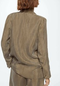 Personne aux cheveux courts marron portant une chemise marron à manches longues et ample, ainsi qu'un pantalon assorti, debout de dos face à la caméra.