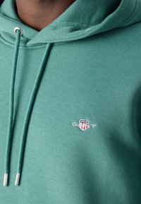 Gros plan d'une personne portant un sweat à capuche vert avec des cordons de serrage et un petit patch logo GANT sur la poitrine.