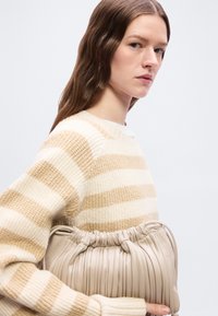 Γυναίκα με μακριά καστανά μαλλιά, ντυμένη με πουλόβερ πλεκτό σε μπεζ και λευκές ρίγες, κρατώντας μια μπεζ πλεκτή δερμάτινη τσάντα με πιέτες εναντίον ενός απλού φόντου.