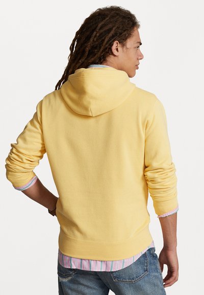 Rumena kapuca s sproščenim krojem in kapuco, nosi se čez sivo in modro črtasto srajco. Ima dolge rokave in rebraste manšete.