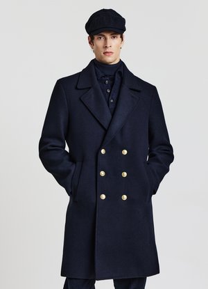 Jeune homme portant un manteau croisé bleu marine avec des boutons dorés et une casquette bleu marine assortie, debout les mains dans les poches.