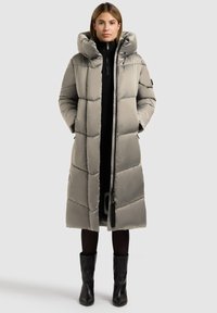 Langer, gepolsterter grauer Mantel mit hohem Kragen und übergroßer Kapuze, ausgestattet mit einem vertikalen Reißverschluss und seitlichen Taschen. Wird über einem schwarzen Rollkragenpullover getragen.