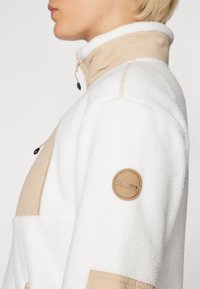 Giacca in pile bianco con dettagli beige, colletto alto e una toppa sul maniche. Presenta materiali a contrasto e un dettaglio con zip.