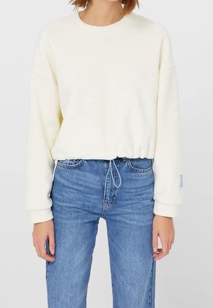 Femme portant un sweat-shirt court à manches longues couleur écru avec un cordon de serrage à l'ourlet et un jean bleu taille haute, debout devant un fond uni.